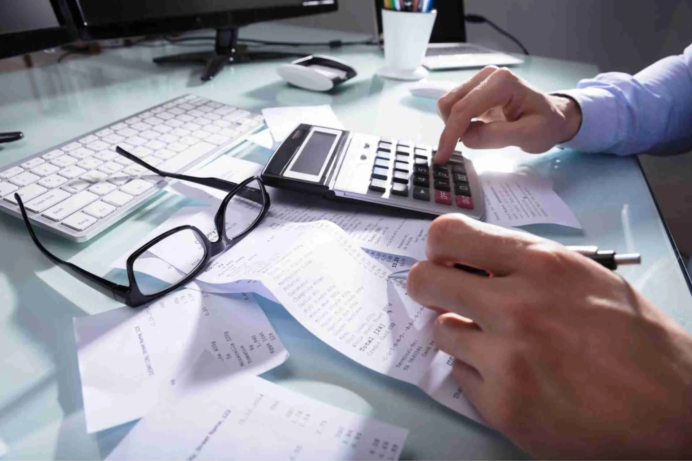 Hands using a calculator with receipts, glasses, and a keyboard on the desk.