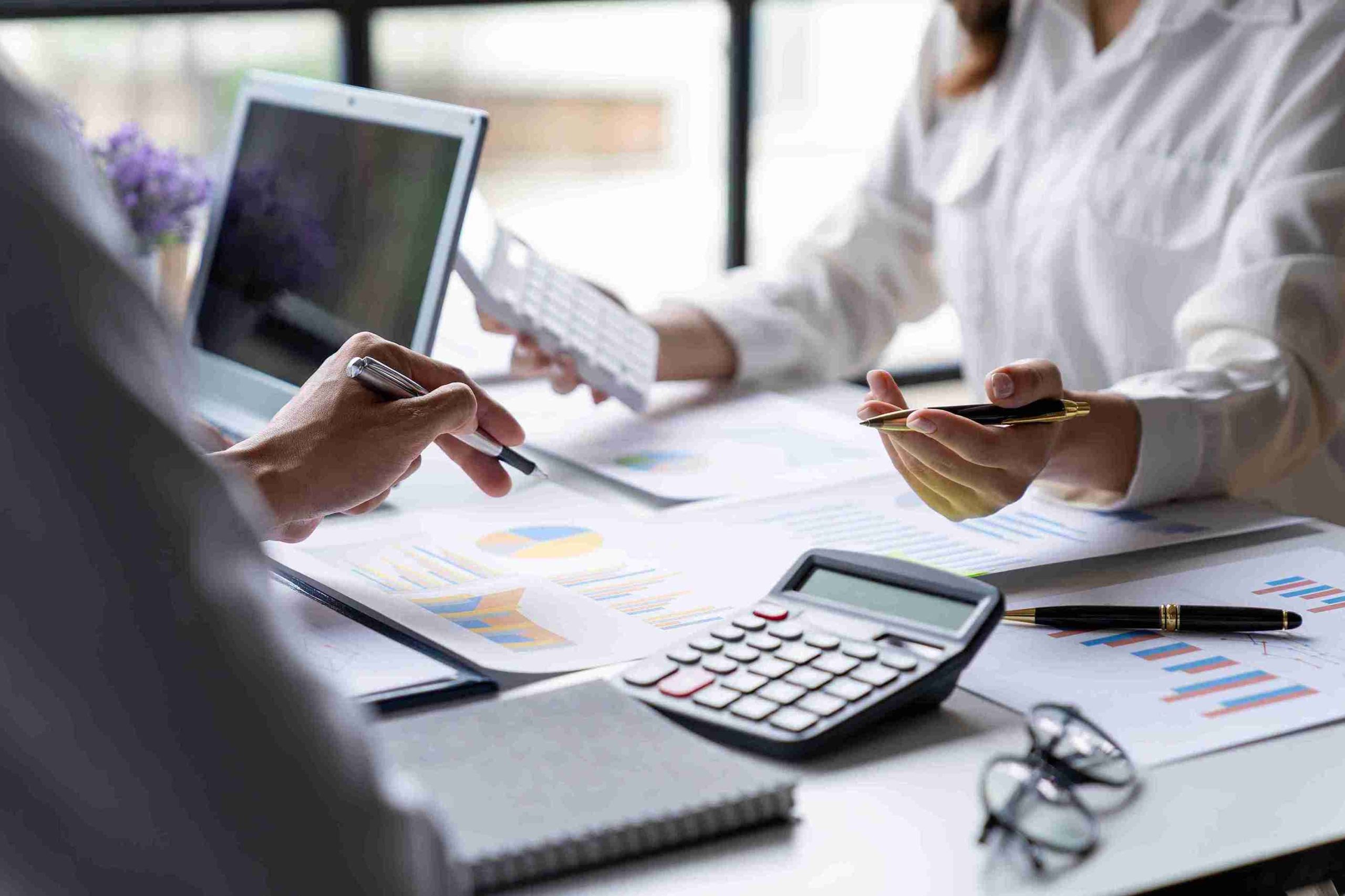 Business professionals collaborating on a laptop, surrounded by papers and documents in a modern office setting.