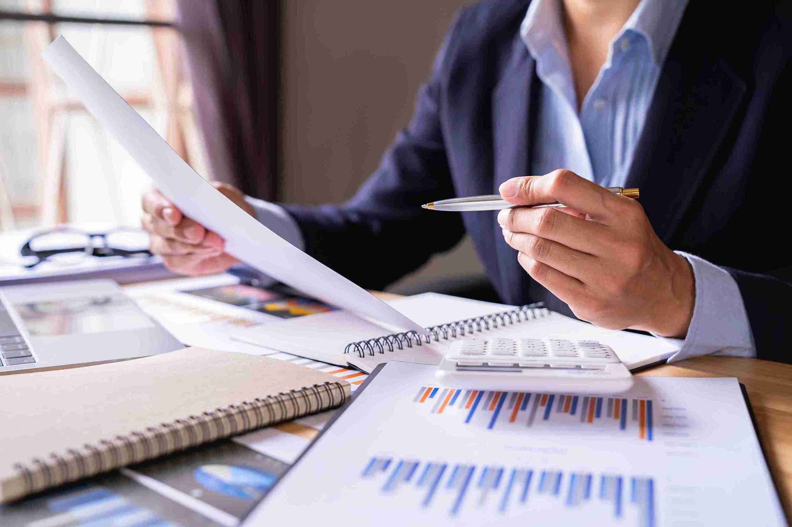 A man in a suit sits at a desk, surrounded by papers and a calculator, focused on his work.