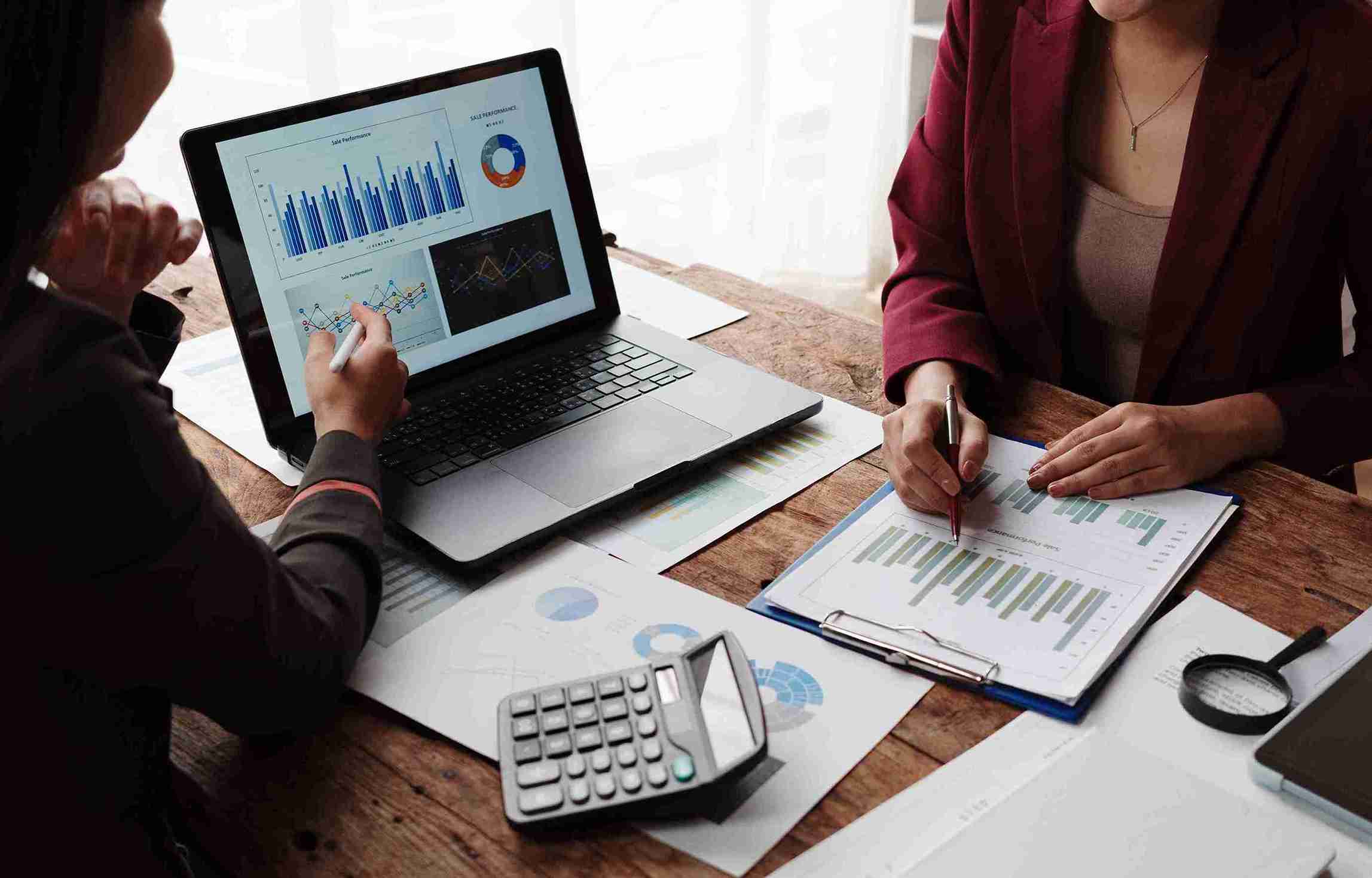Two business professionals collaborate at a table, reviewing documents and working on a laptop together.