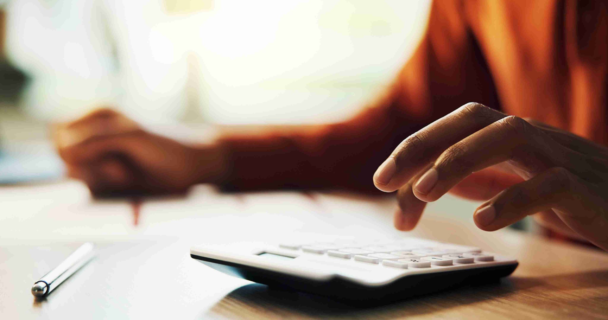A person focused on a calculator while sitting at a desk, surrounded by papers and a pen.