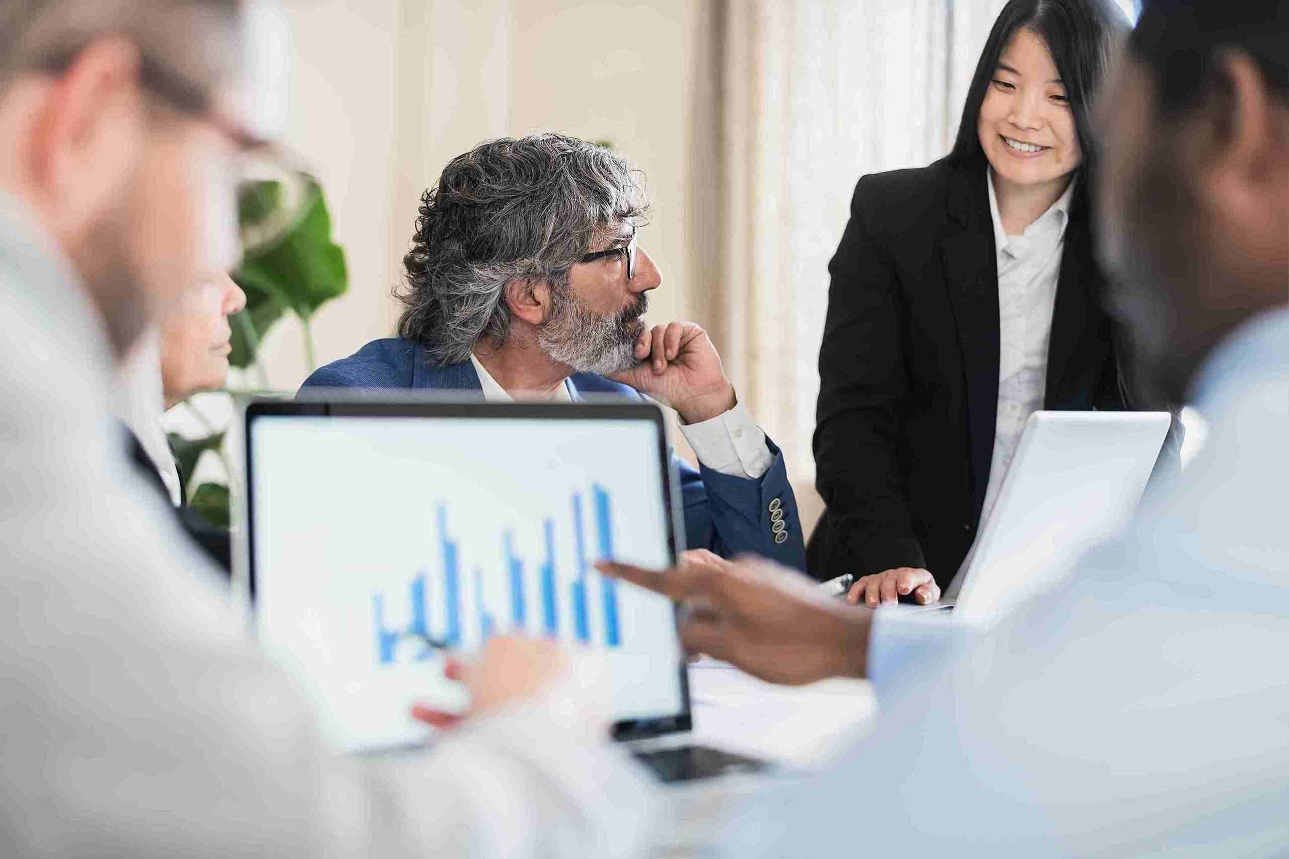 A diverse group of people collaborating in a meeting room, focused on a laptop in the center of the table.