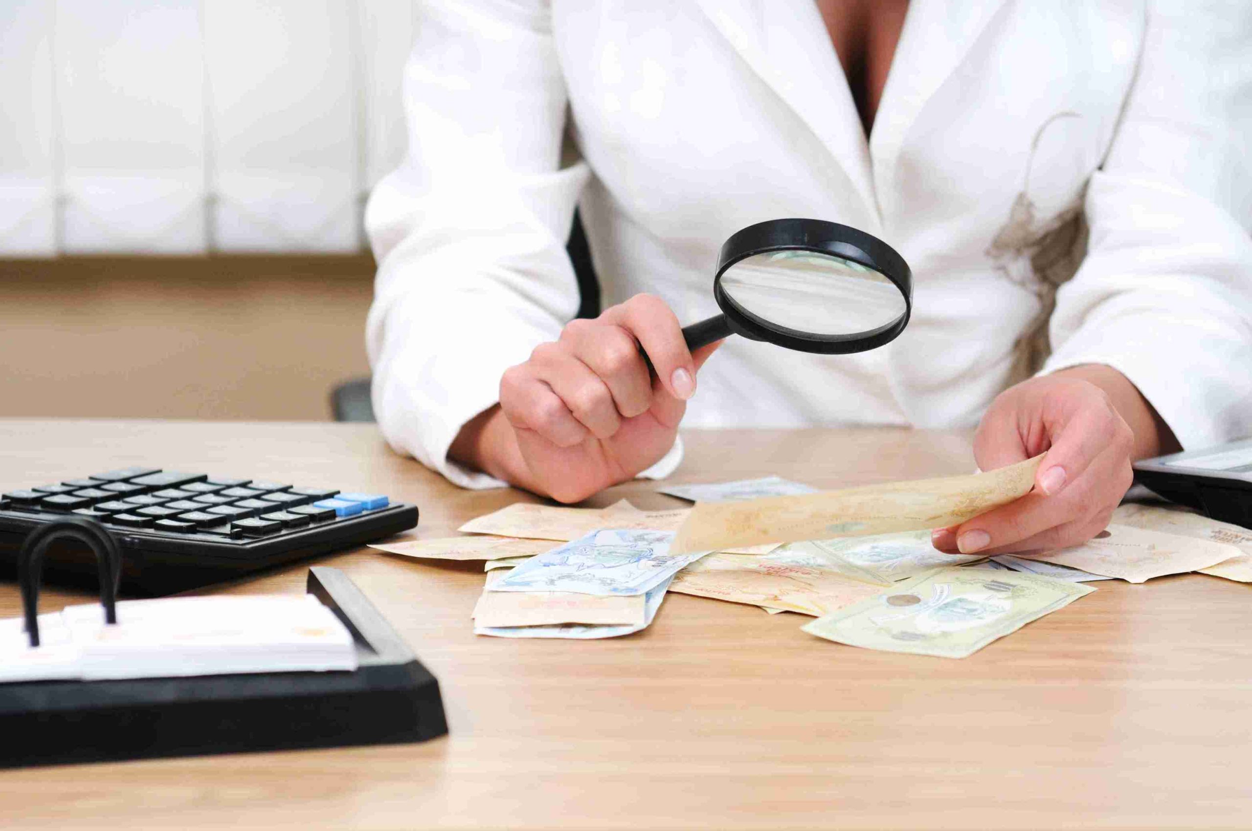 A woman in a white coat examines a pile of money with a magnifying glass, focusing intently on the details.