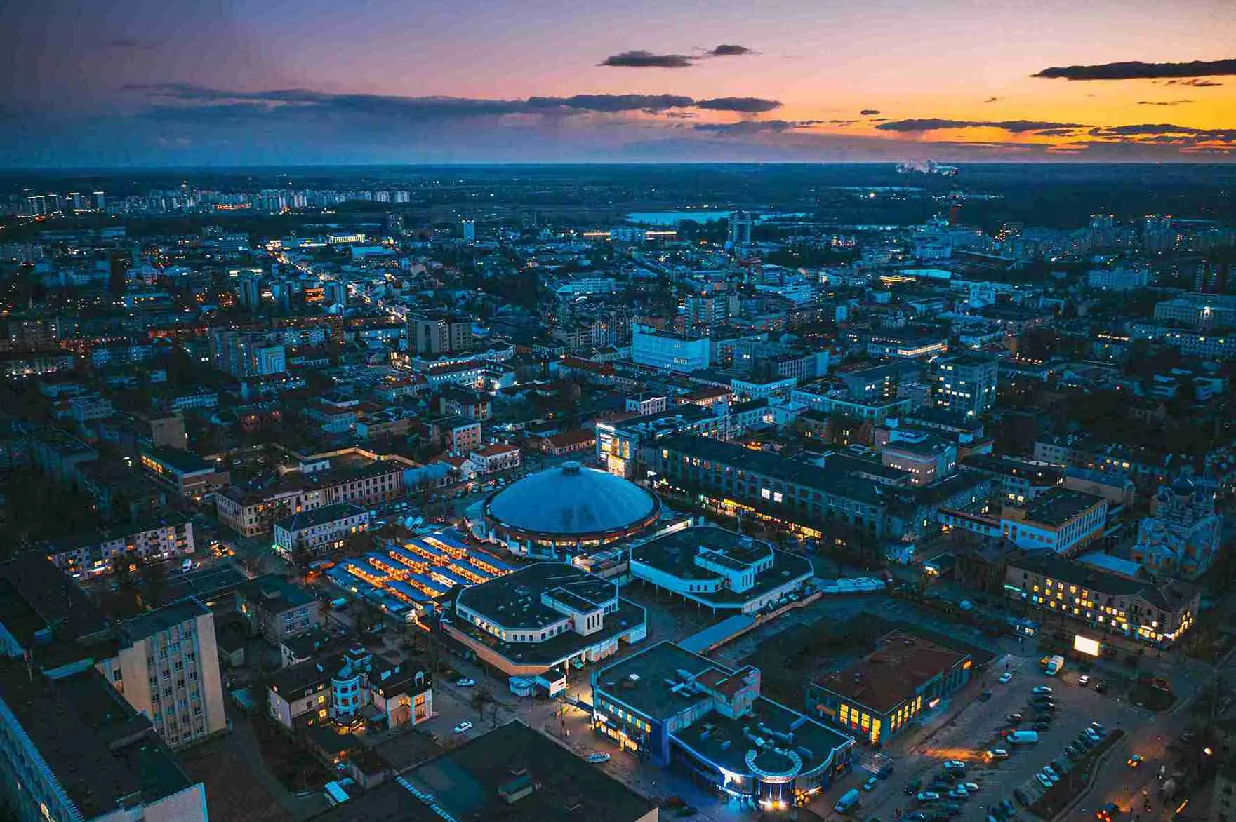 Kiev skyline at dusk, showcasing illuminated buildings and a vibrant sky transitioning from orange to deep blue.